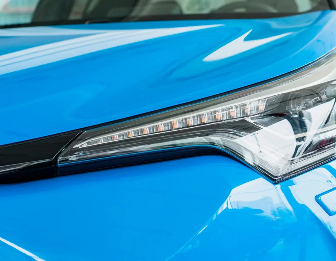 A close-up of a blue car’s hood and headlight, showcasing an exceptionally shiny and reflective surface. The polished finish highlights the car’s vibrant color and flawless shine, with light reflecting off the smooth, glossy paint.