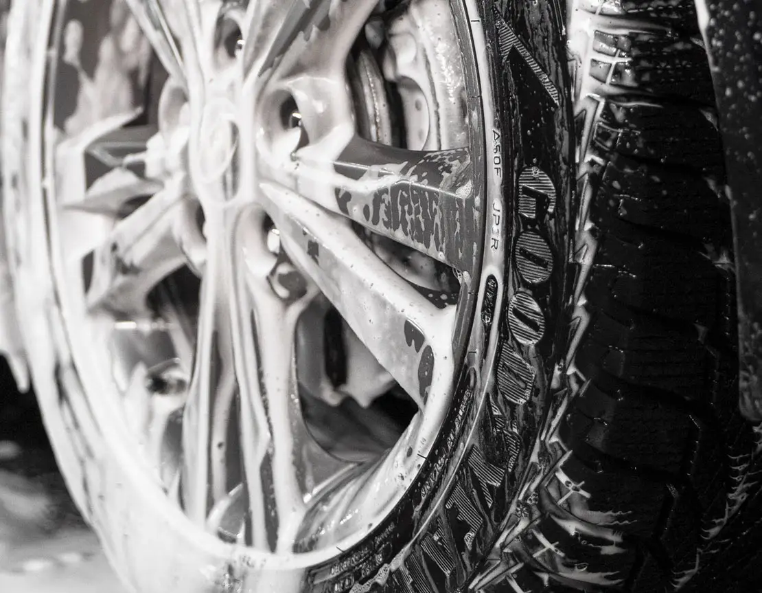 A tire covered in thick, foamy suds, with soap bubbles clinging to its surface as it is being thoroughly cleaned, highlighting the detailed attention given to removing dirt and grime from the tire.