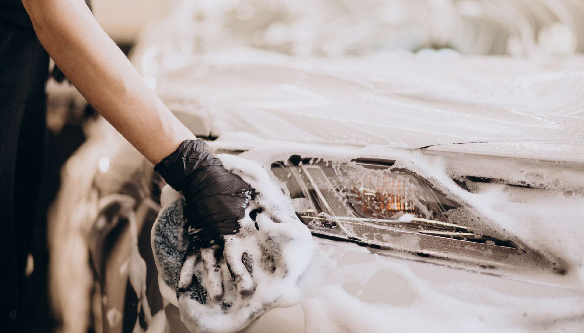 Someone using a sponge to wash their car, covered in suds, with foam lathered on the car's surface as they clean it.