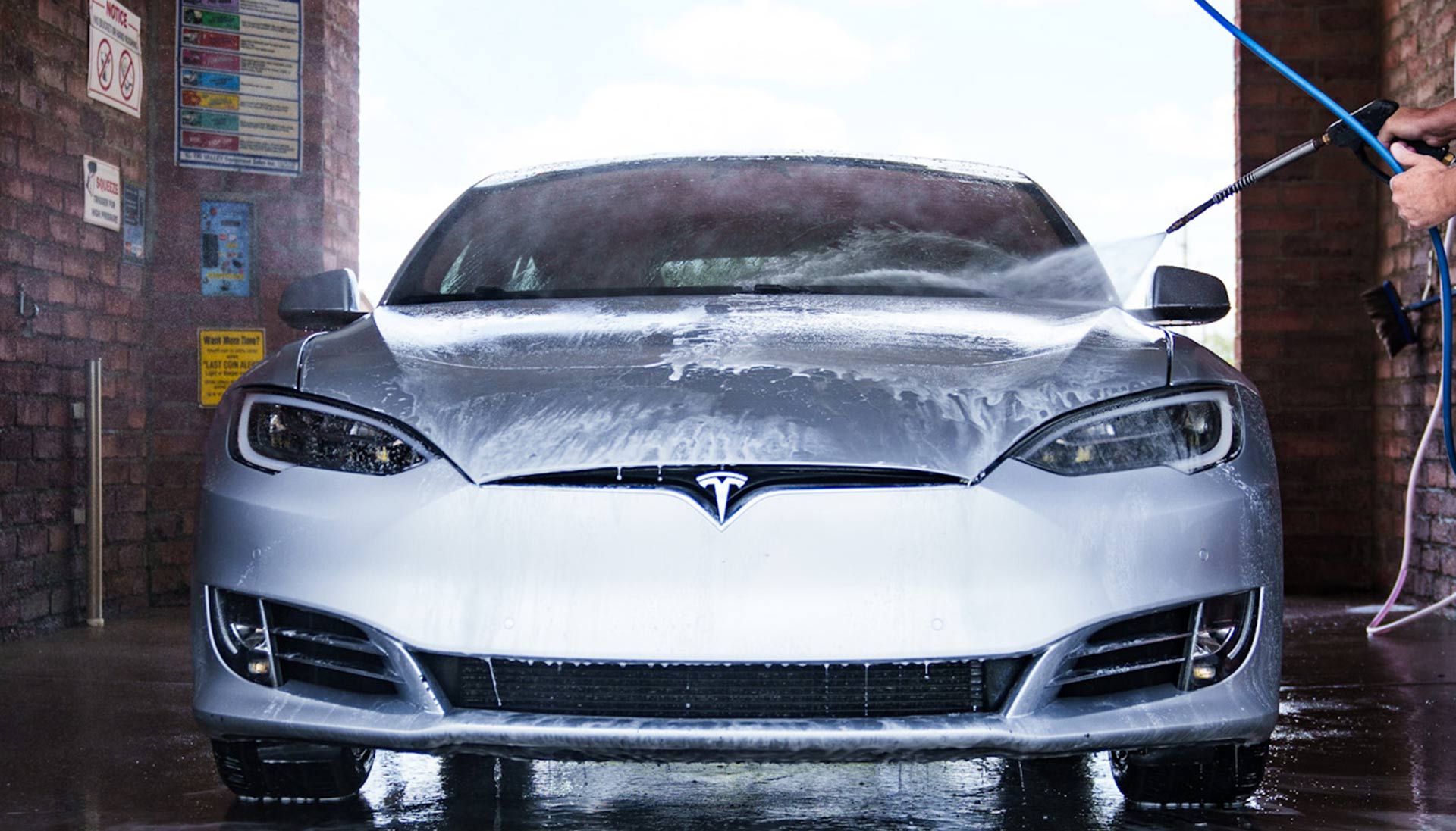 A grey Tesla being washed in a self-service car wash bay, covered with suds and foam, with the vehicle thoroughly coated as it undergoes cleaning.